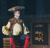 Falstaff, with protruding belly, sits on a crate and holds a tankard in his left hand, his right hand held out as if in regal posture with his mouth puckered in great pronouncement: he has a pillow on his head, and he's wearing his normal outfit of buckskin vest over yellow shirt, blue pants, and bright redk, knee-high boots. A banner of three royal lions hang on a brick wall in the background.